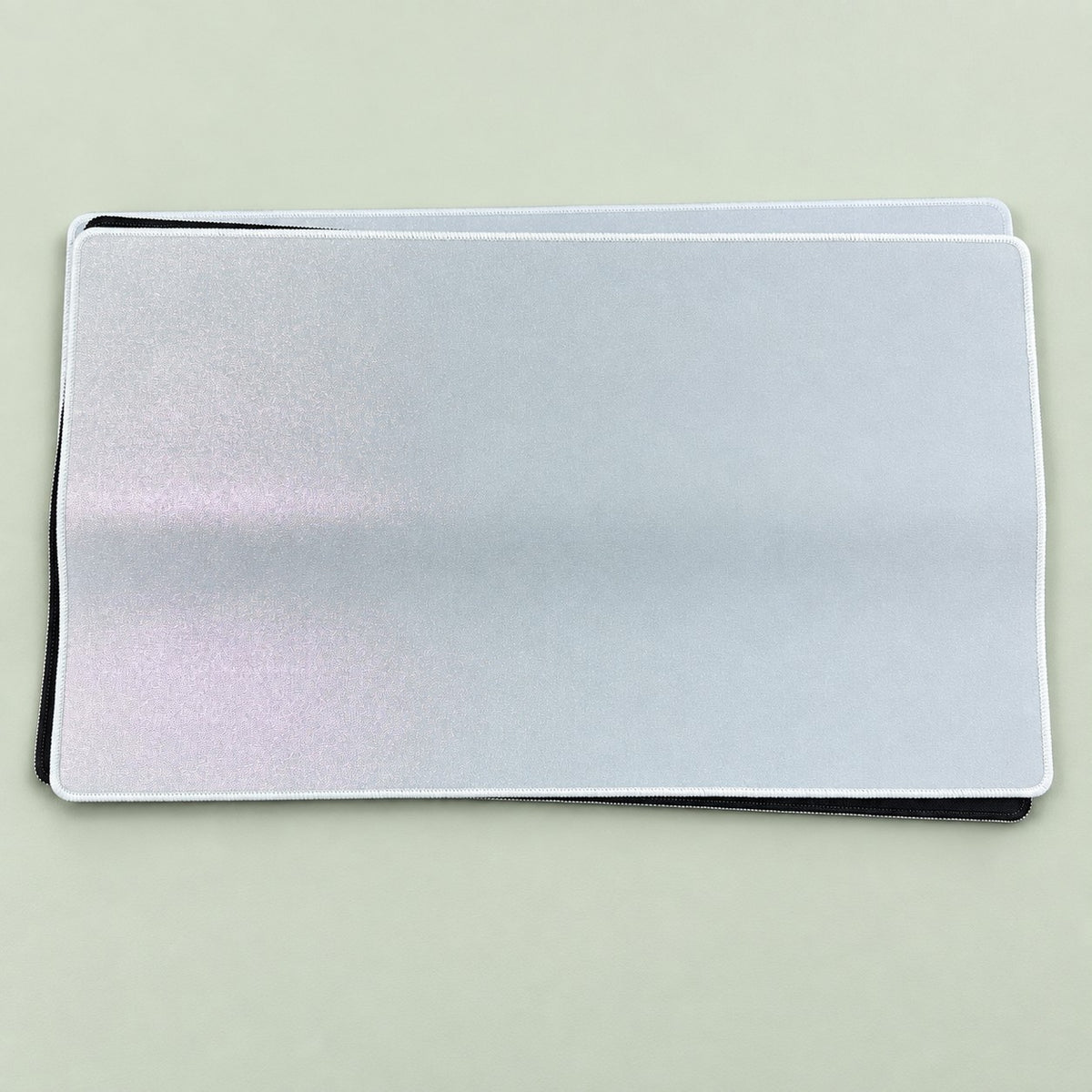 Stack of blank holographic sublimation TCG playmats showing white holofoil top and black rubber base